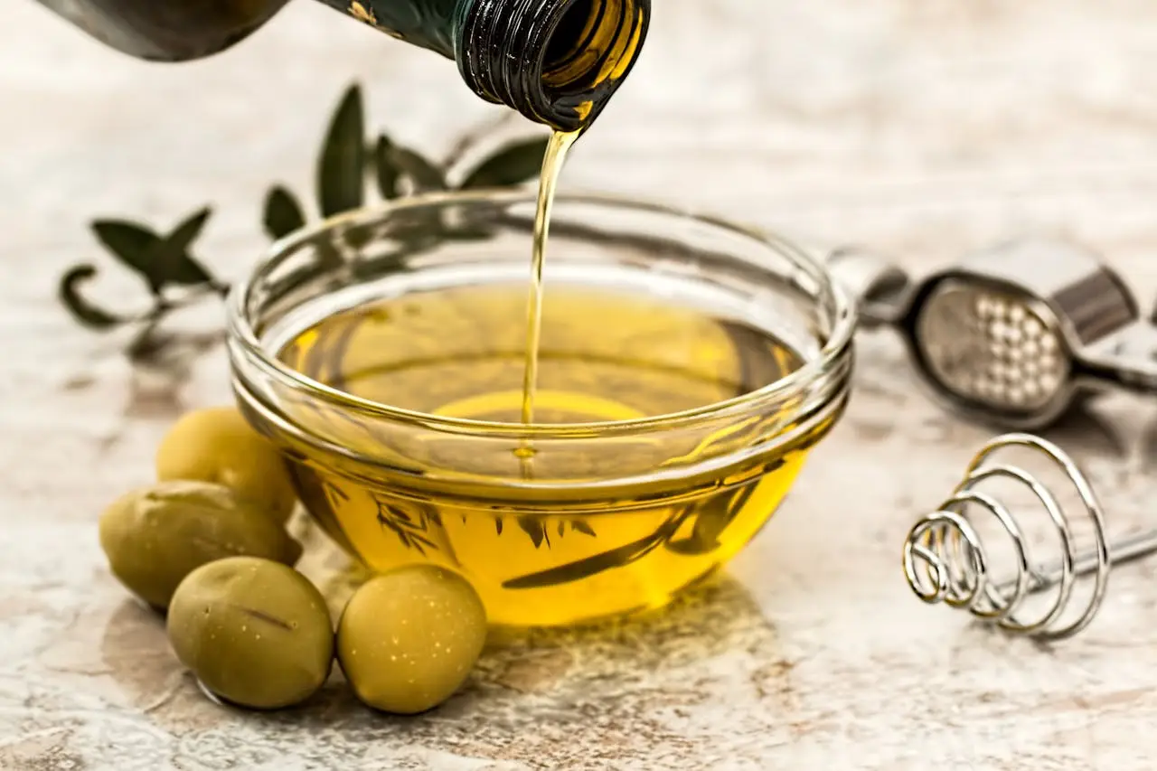 who-we-are Close-up of olive oil being poured into a glass bowl surrounded by fresh olives and kitchen tools.